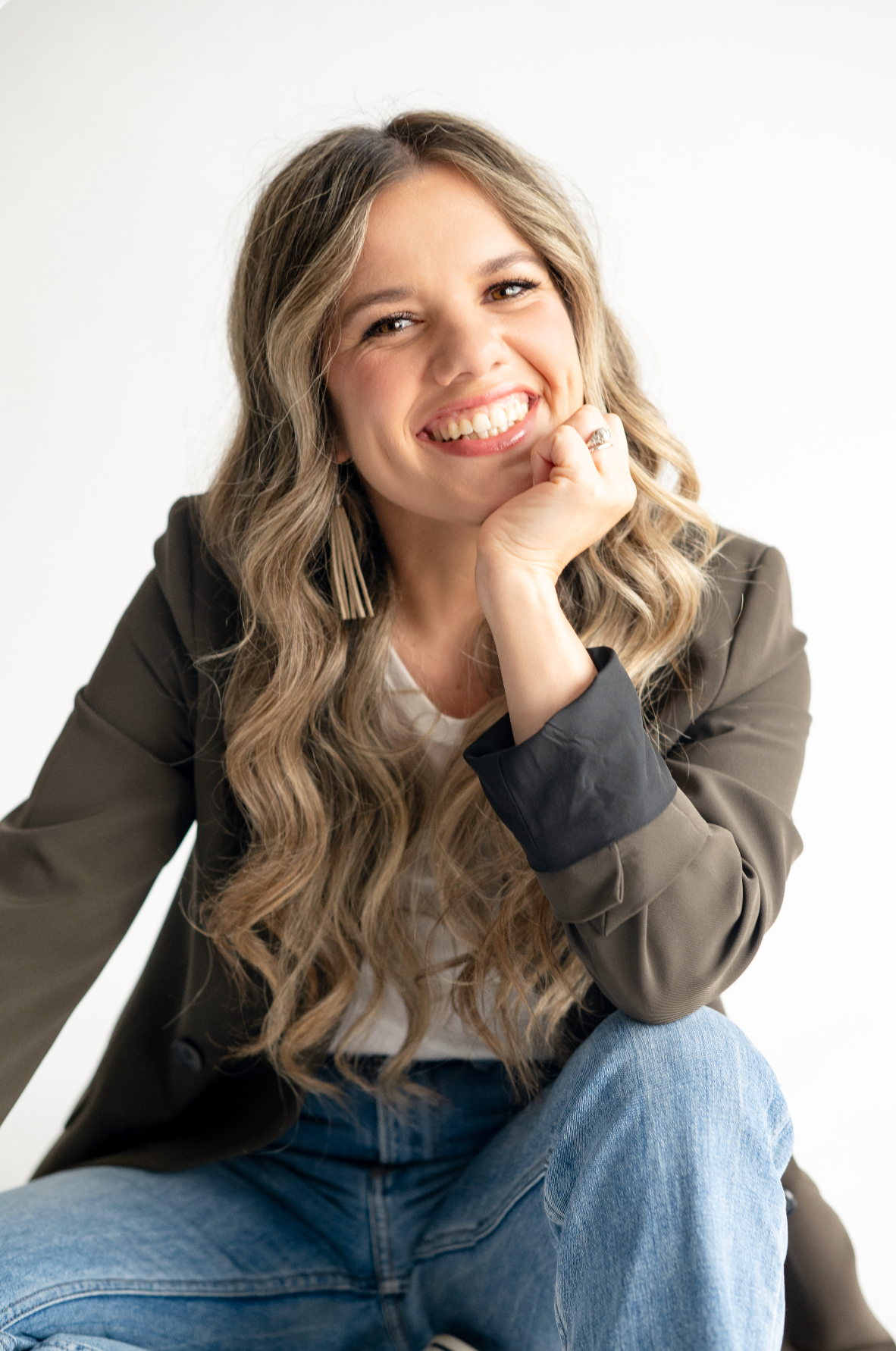 Professional studio portrait of someone in a blazer and jeans with long wavy hair sitting casually against white background.