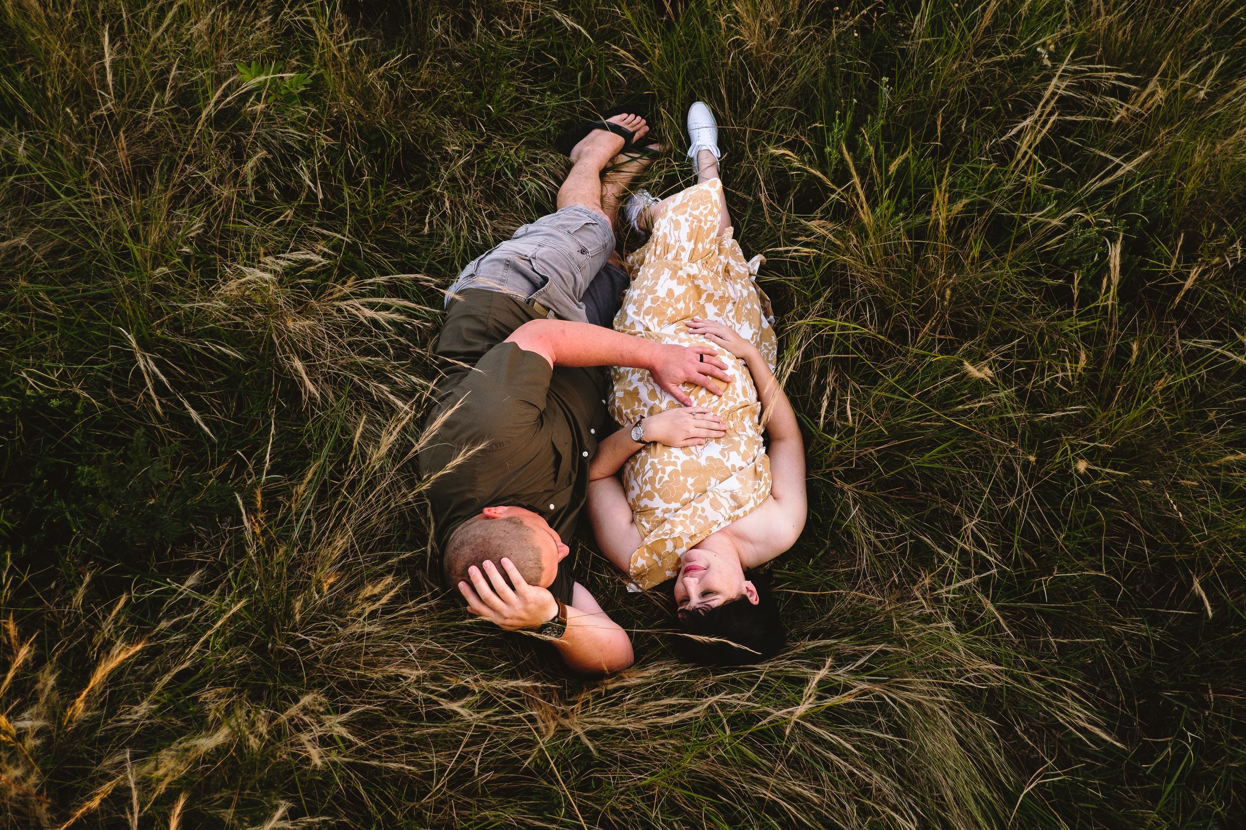Maternity couple lying in long grass, sharing a quiet moment of connection
