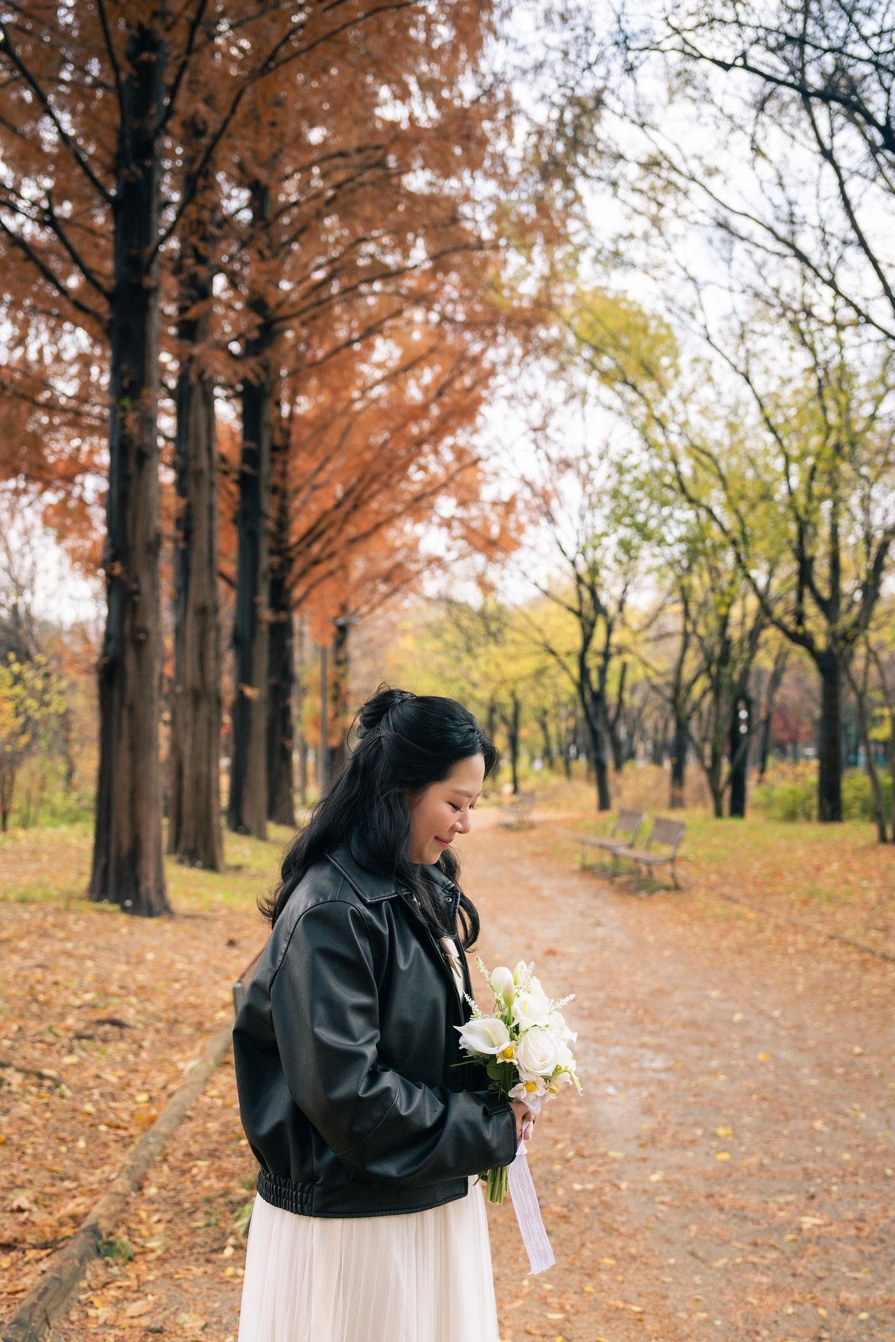 A person in a black jacket holds white flowers while walking along a tree-lined autumn path with fallen leaves.