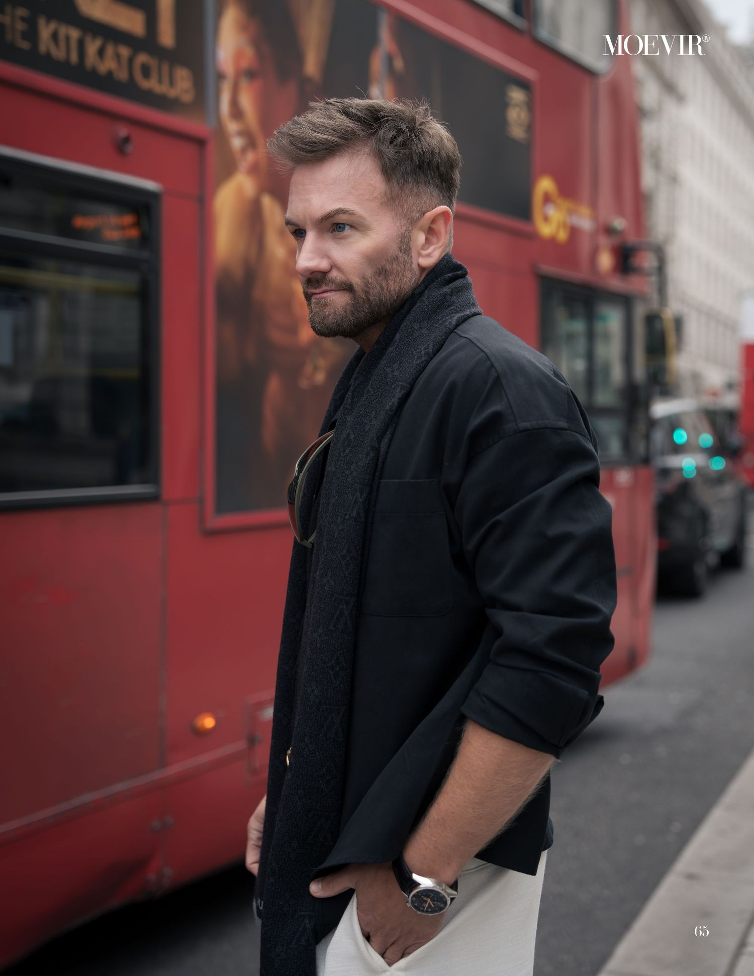 A person in a black coat stands on a city street with a red storefront in the background.