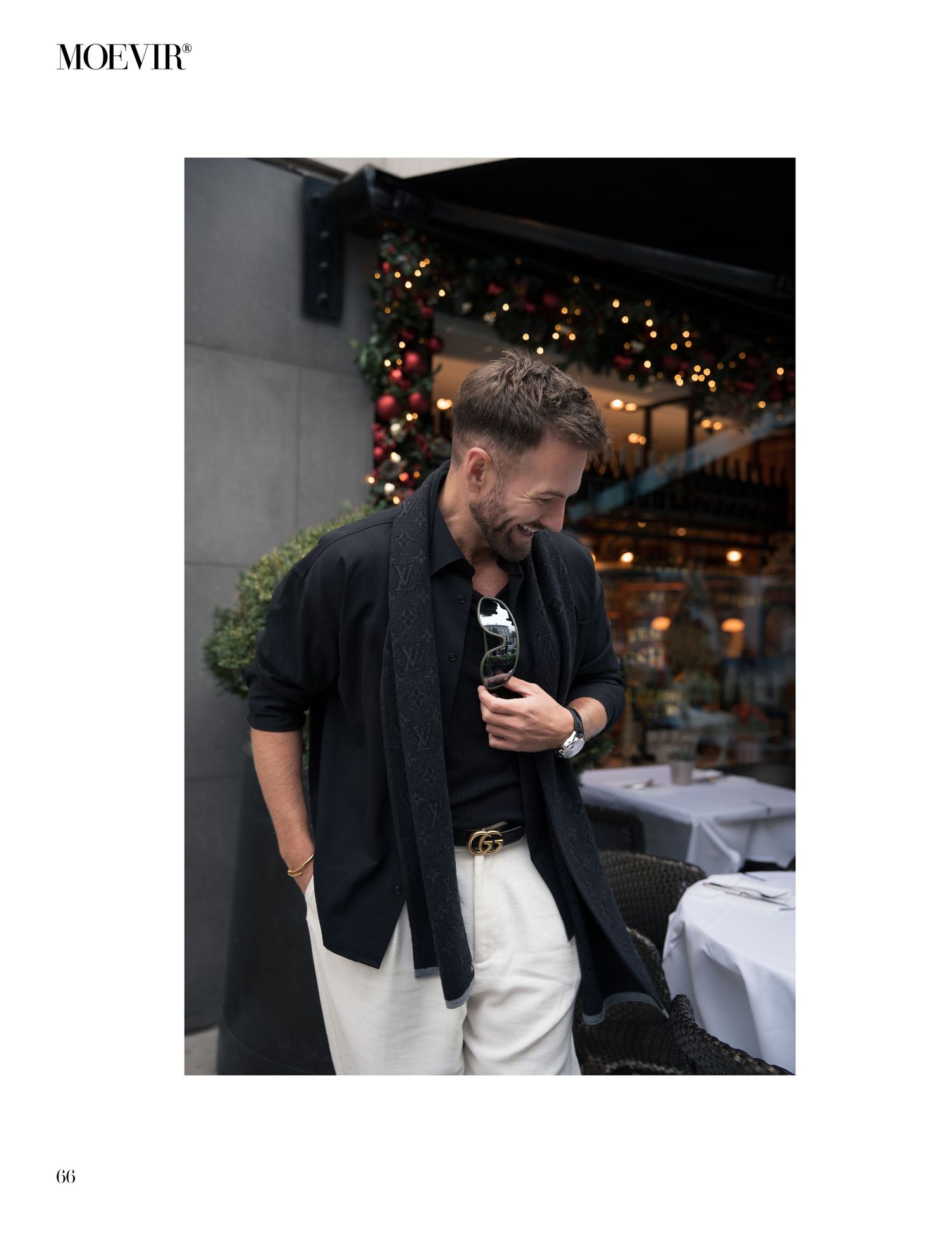 A person in a black blazer and white pants stands outside a restaurant with holiday decorations and twinkling lights.