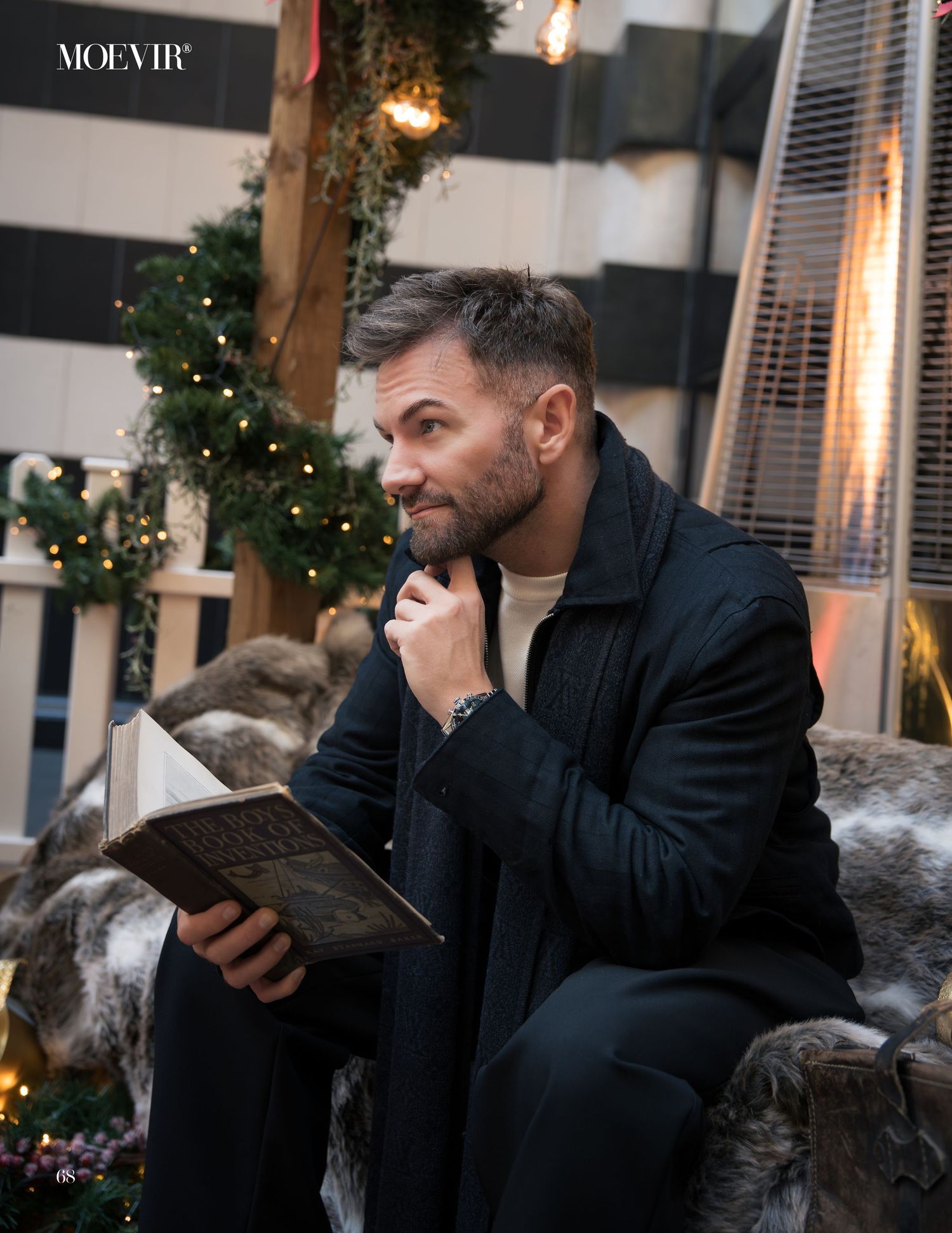 A person in a dark suit sits on a couch reading a book in a room decorated with Christmas lights and greenery.