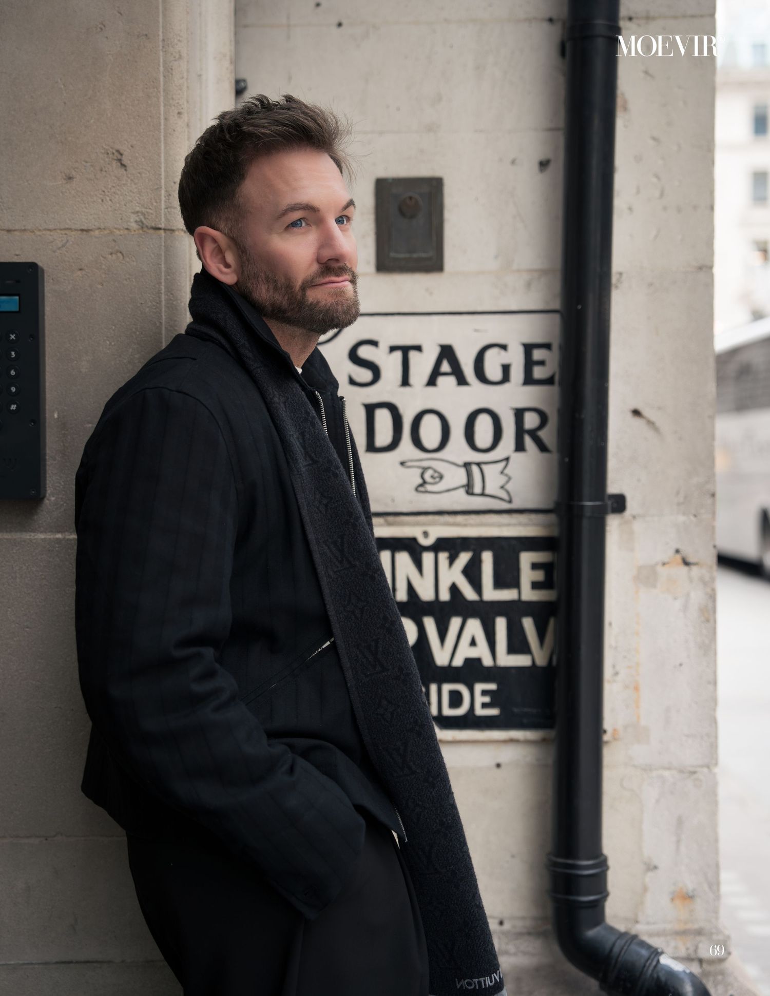 A person in a black coat stands against a concrete wall with stage door signage in an industrial setting.