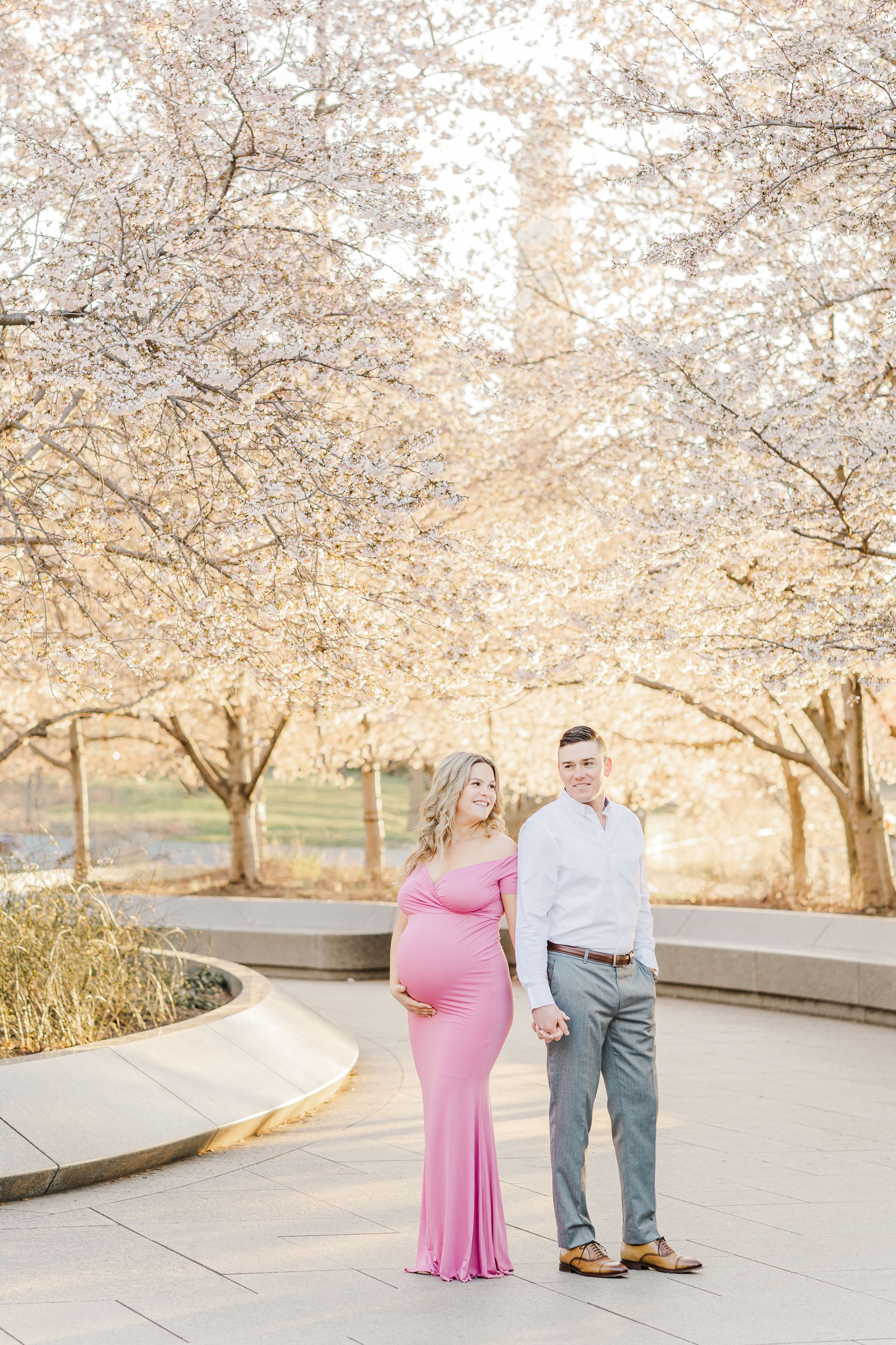 Expecting couple poses under blooming cherry blossom trees in springtime.