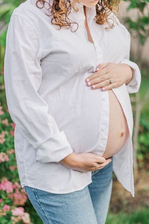 Woman in white button-down shirt showing her pregnant belly in garden setting.