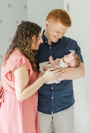 A couple in pink and navy blue clothing share a tender moment while holding their newborn baby.