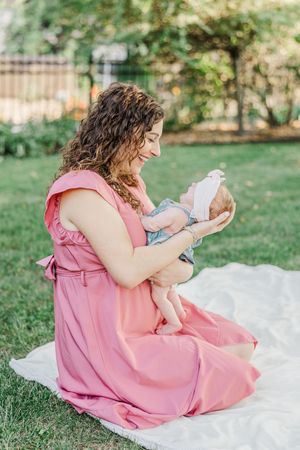 Mother in pink dress sitting on white blanket outdoors holding newborn baby.