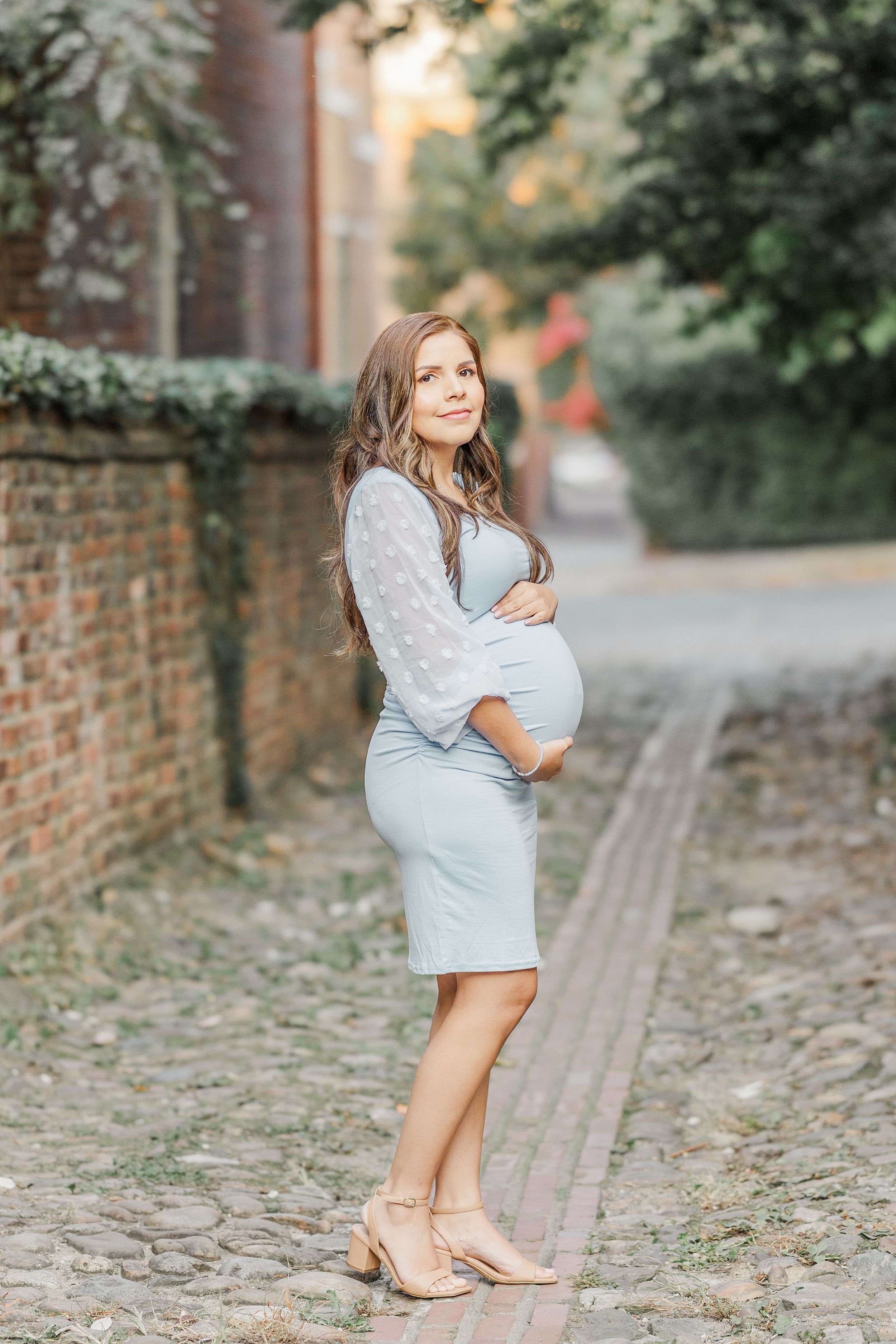 Expectant mother in a light blue dress and sweater poses on a cobblestone path near a brick wall.