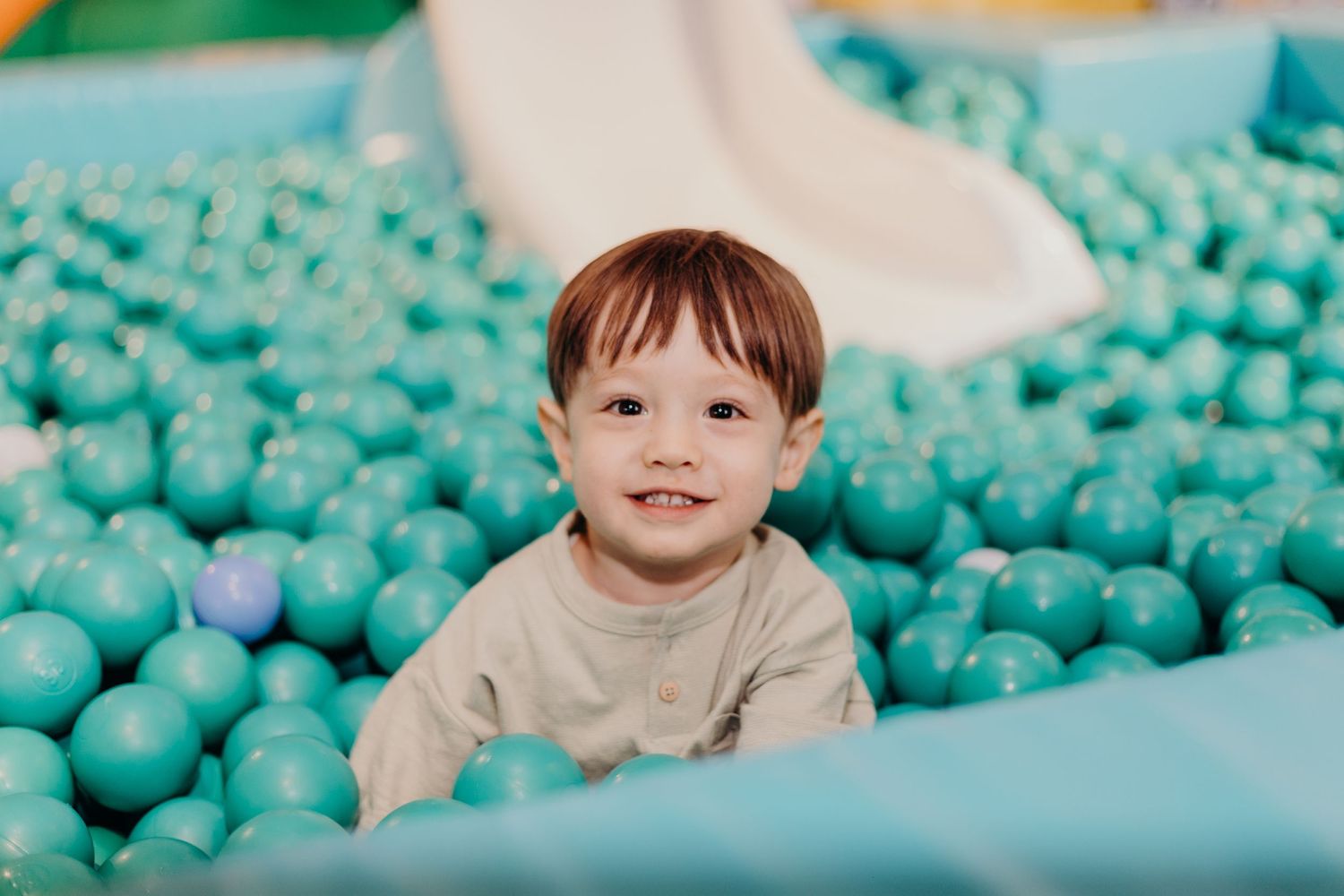 A child plays in a pool filled with turquoise blue plastic balls at an indoor playground attraction.
