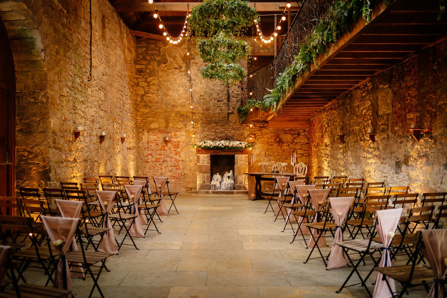 Rustic stone barn venue set up for wedding ceremony with wooden chairs and string lights along stone walls.