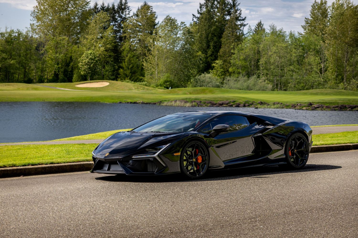 A sleek black Lamborghini sports car parked on a road beside a scenic golf course with trees and a lake.