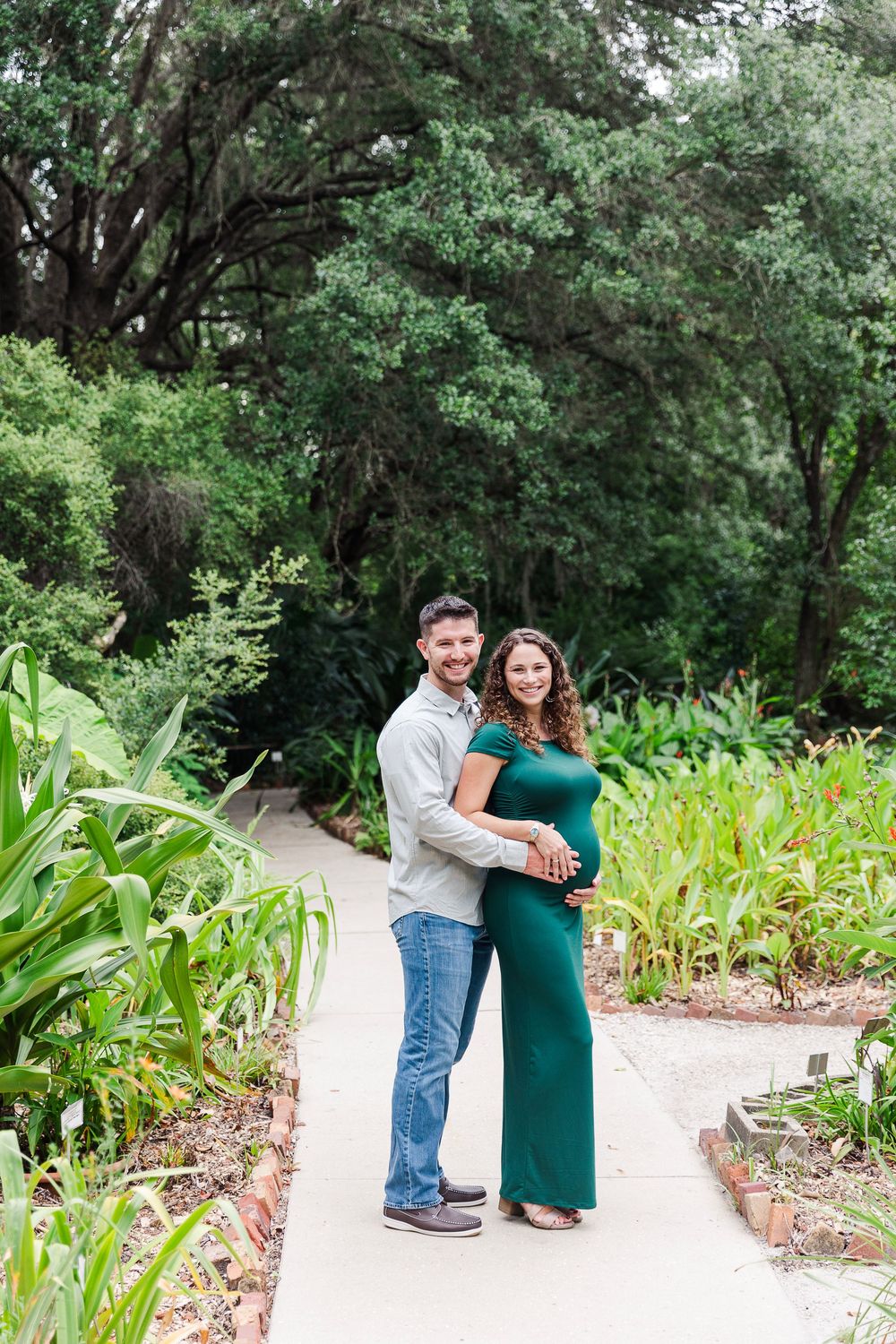 A couple embraces outdoors in a lush garden setting with green foliage and a winding path.