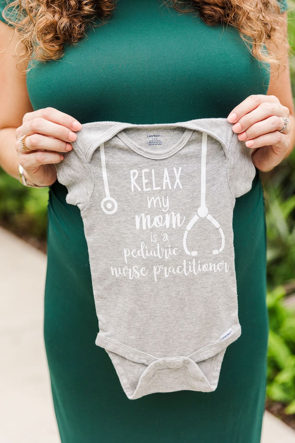 A person in a green dress holds up small gray baby onesies with cute text designs.