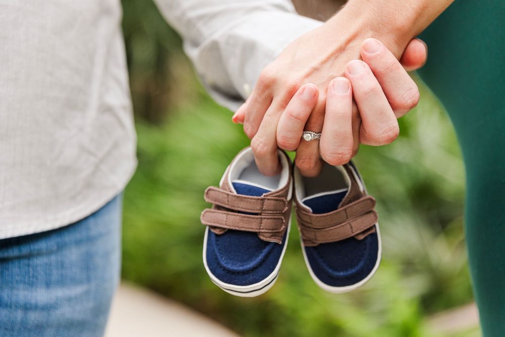 A row of tiny blue baby shoes being held up against a blurred green outdoor background.
