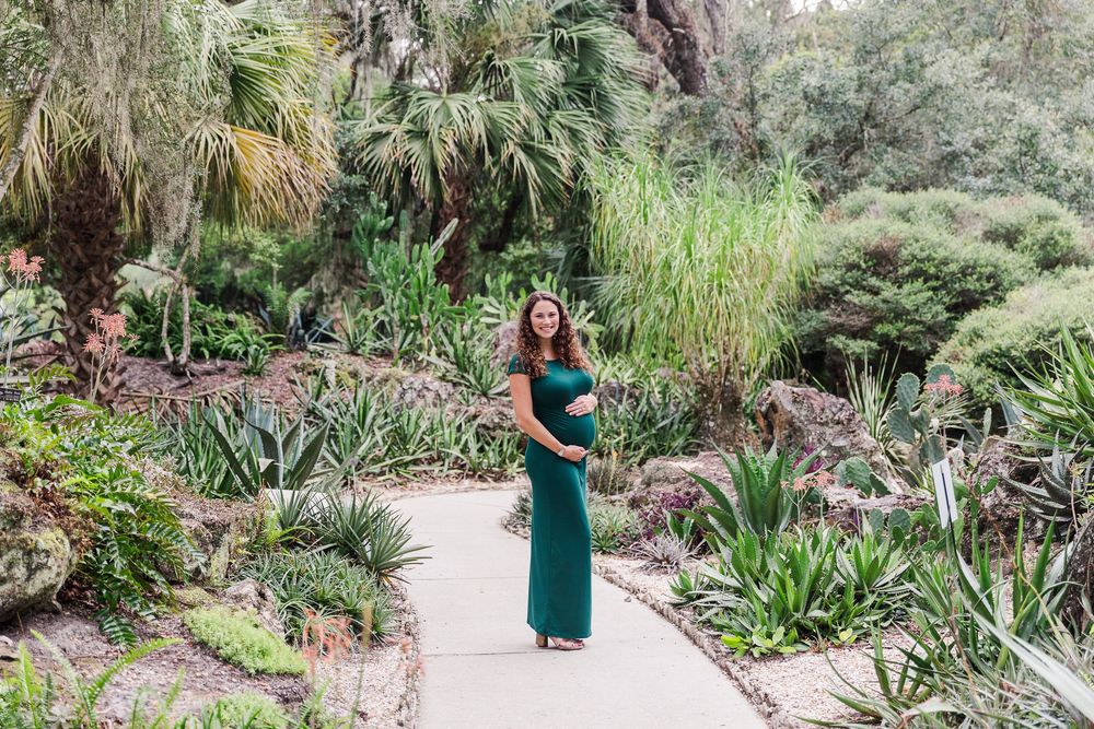 A person in a green dress poses in a lush botanical garden setting surrounded by tropical plants and foliage.
