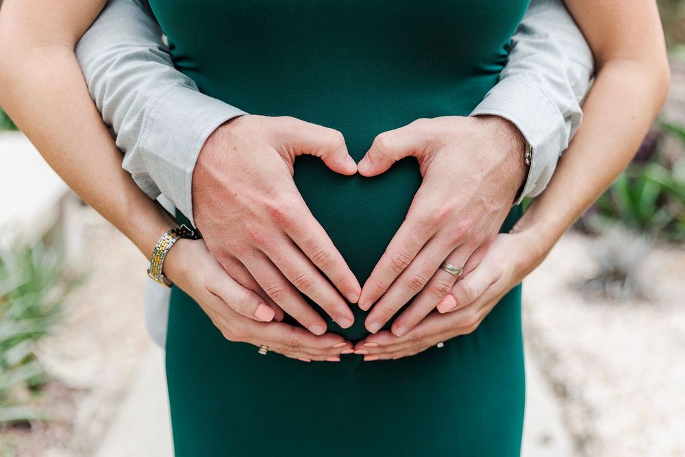 Hands forming a heart shape over a green dress during a maternity photoshoot outdoors.