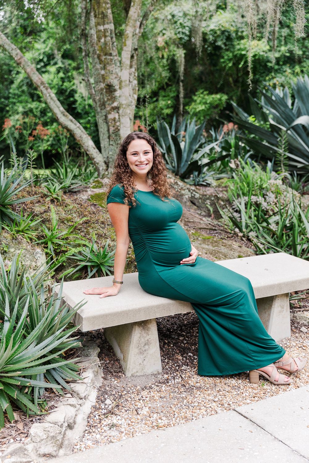 Pregnant woman in emerald green dress sitting on stone bench in lush garden setting.