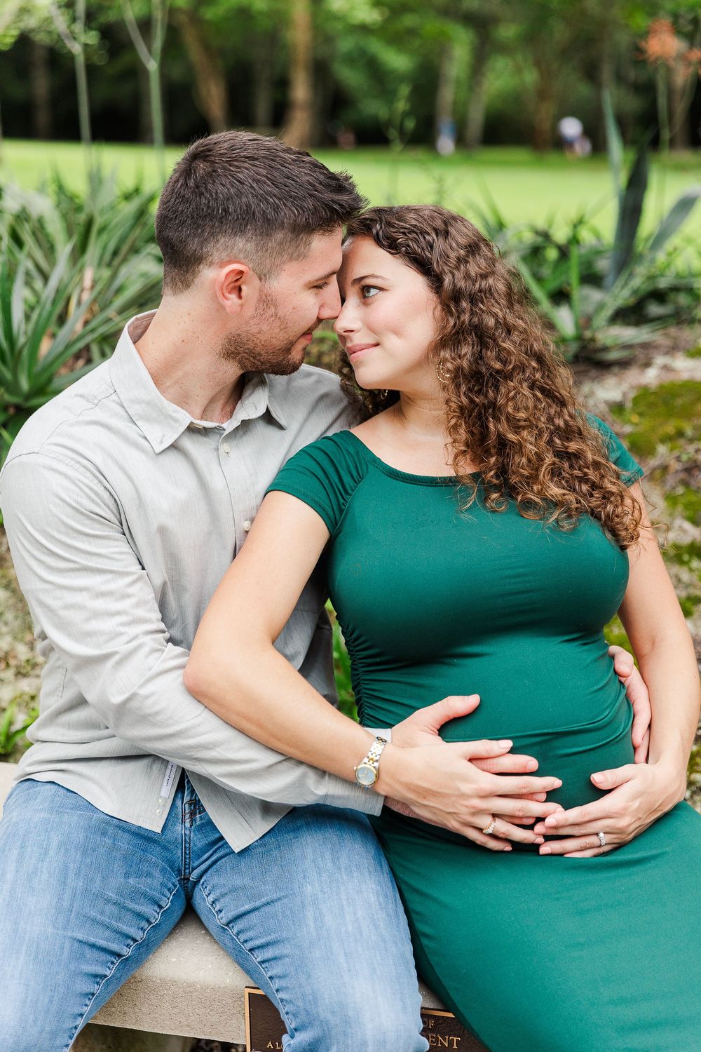 A romantic maternity photoshoot in a garden setting shows a couple in casual attire embracing tenderly.