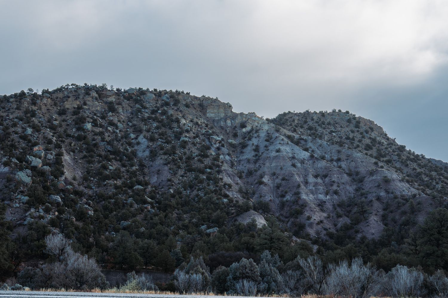 Rocky, tree-dotted mountains under a cloudy sky, with muted colors and rugged terrain creating a calm, natural landscape scene.