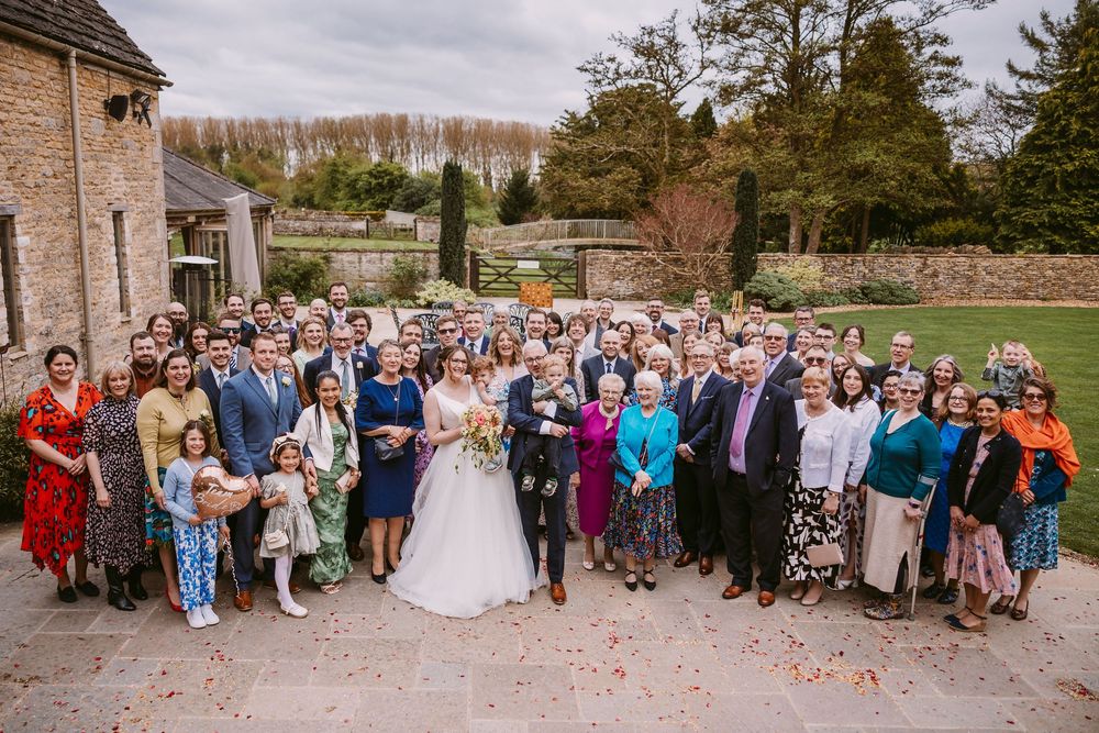 Large wedding party gathered outside historic stone building for group photo on sunny autumn day.