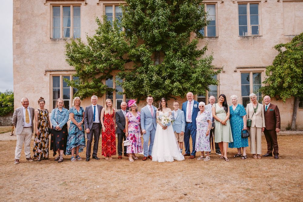 Wedding guests and couple pose in front of stone manor house during outdoor celebration.