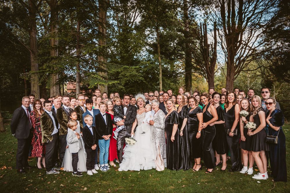 Large wedding party poses together outdoors on a sunny day in front of trees and greenery.