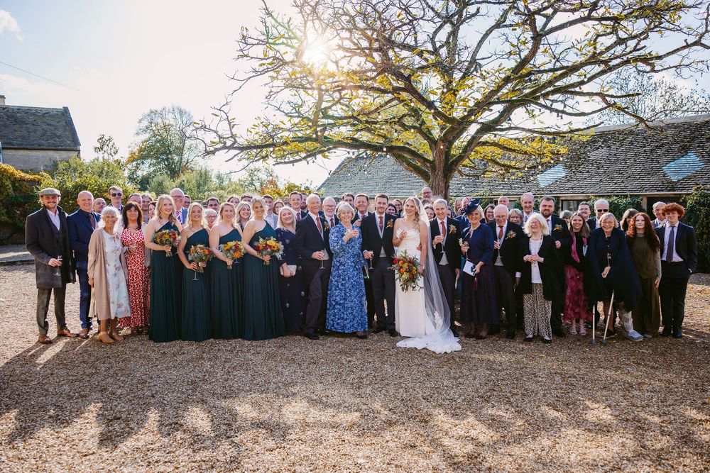 A panoramic outdoor wedding photo showing a large group of guests and wedding party gathered under trees at sunset.