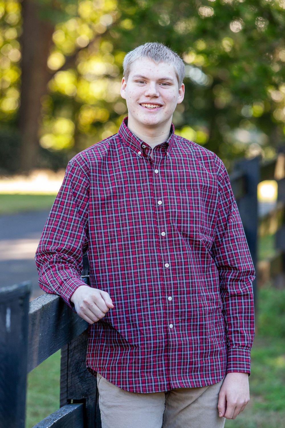 Young man in red plaid shirt leaning on black fence smiling outdoors in fall setting.