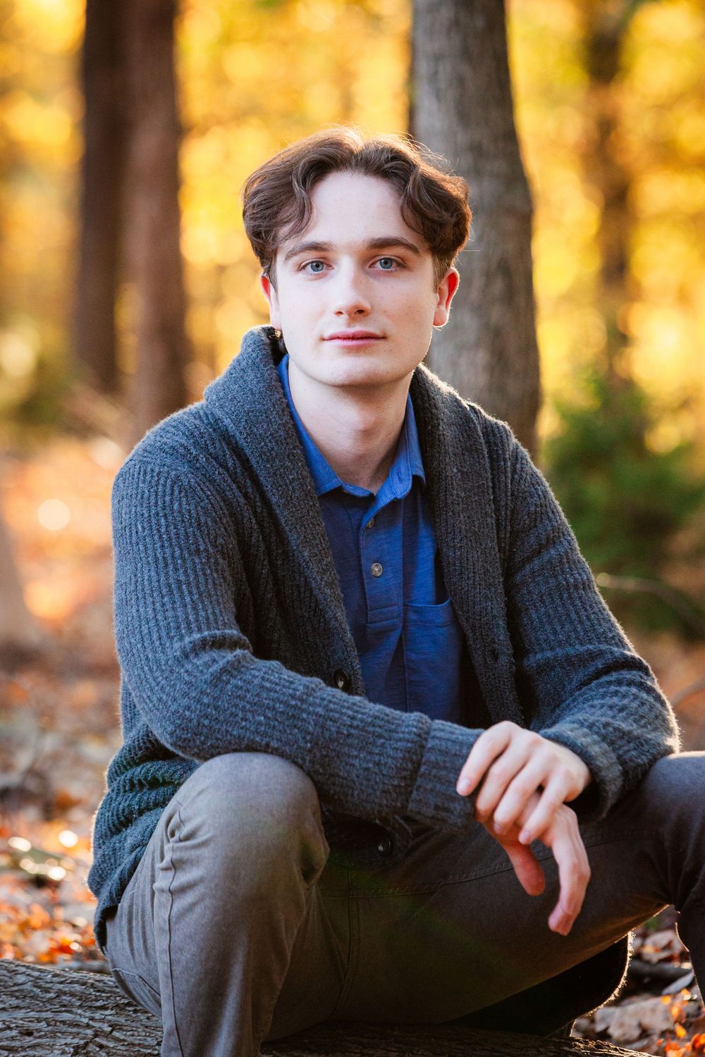Young man in gray cardigan and blue shirt sitting on log in sunny autumn forest.