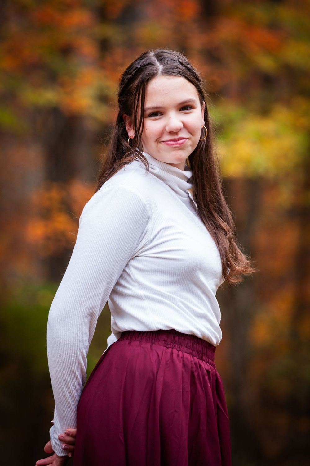 Young woman in white turtleneck and burgundy skirt posing in colorful autumn forest.