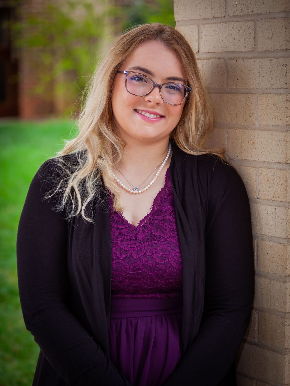 Young blonde woman wearing glasses and pearl necklace posing against a brick wall outdoors.