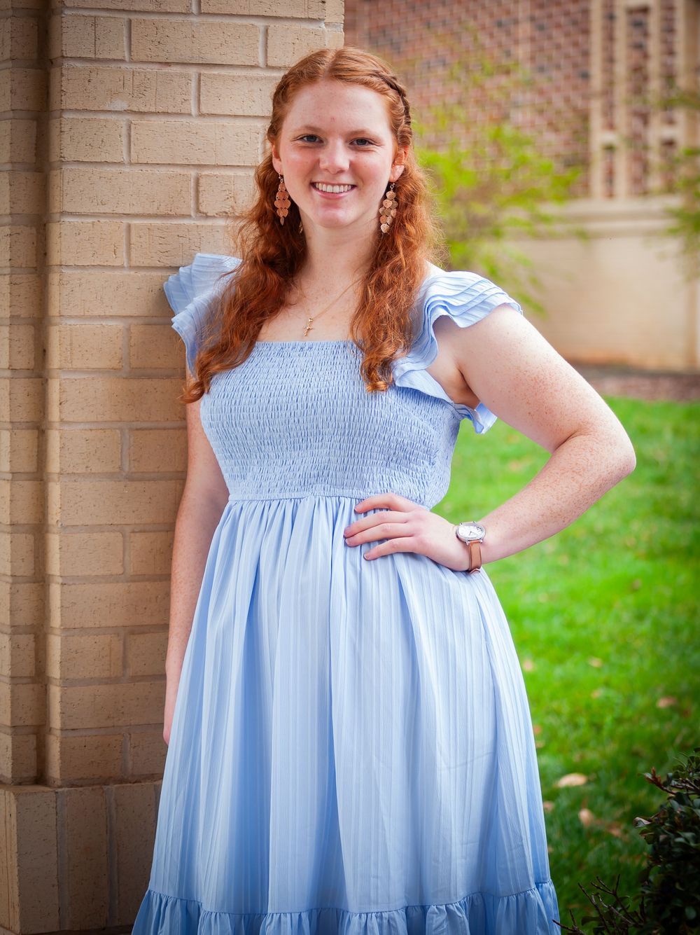 Smiling redhead young woman in a light blue smocked dress posing by a brick wall.