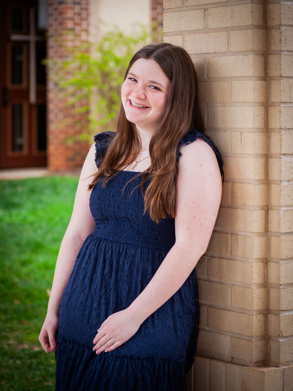 Cheerful brunette young woman in a navy blue dress leaning against a brick wall outdoors.