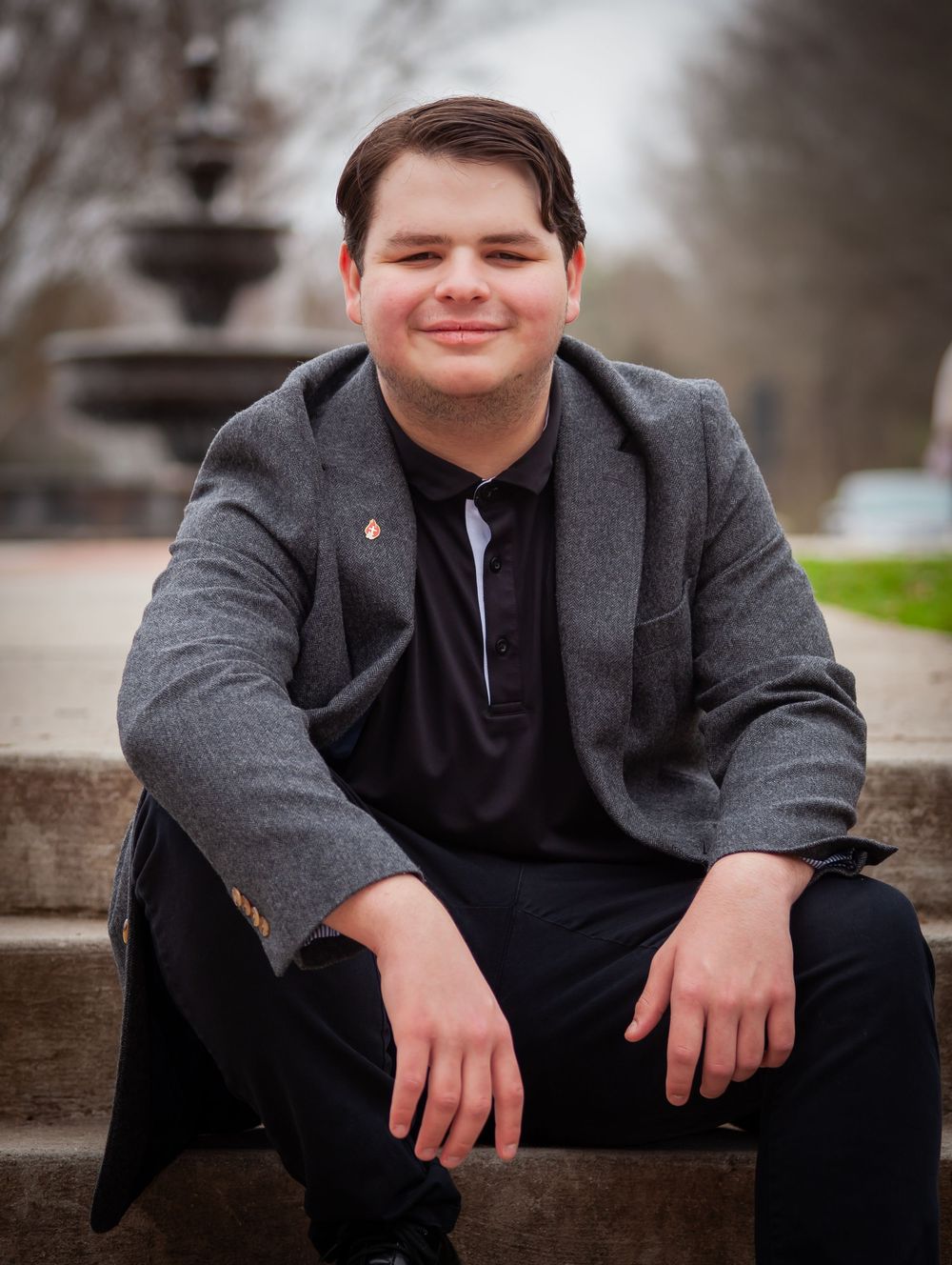 Smiling young man in a gray blazer sitting on outdoor steps near a fountain.