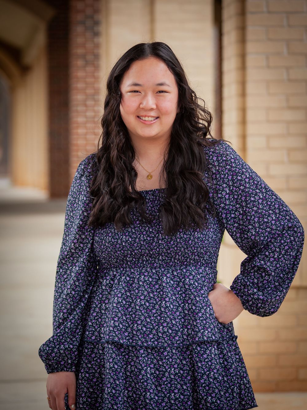 Smiling young Asian woman in a floral navy dress posing confidently by a brick wall.