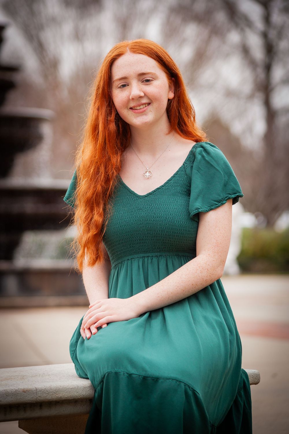 Young woman with long red hair wearing a green dress seated near an outdoor fountain.