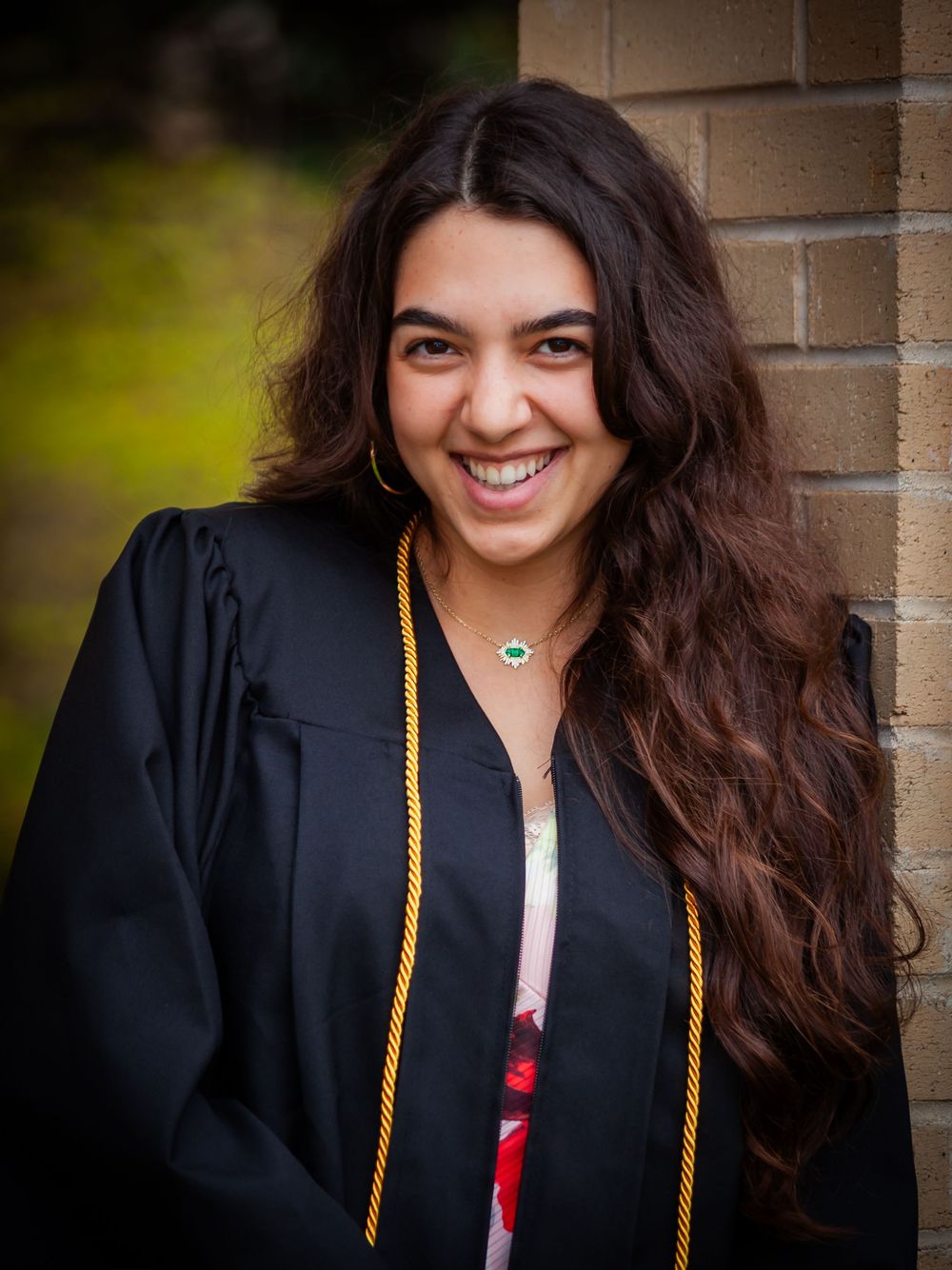 Smiling young woman in graduation cap and gown with gold honor cords posing outdoors.