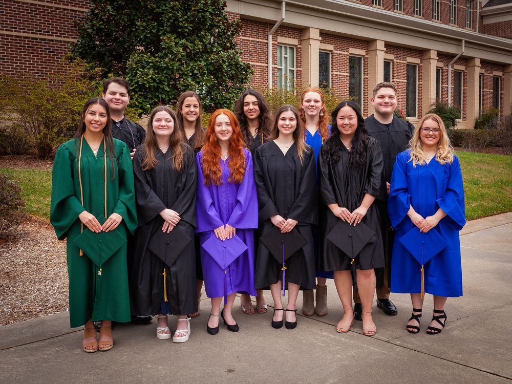 Group of graduates in colorful caps and gowns posing outside brick university building.