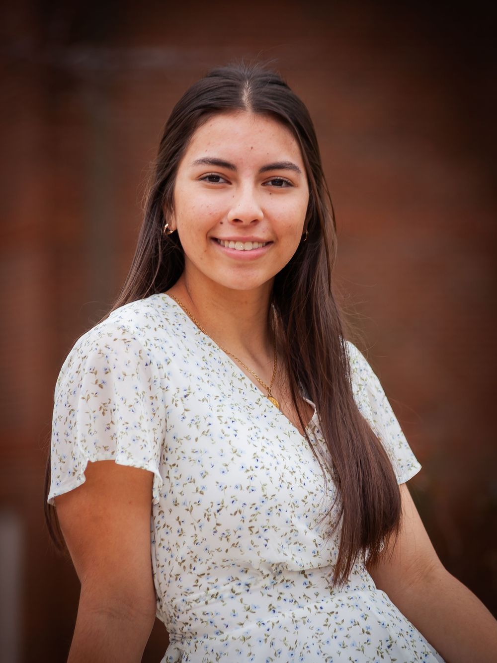 Young woman in floral white dress smiling outdoors against brick wall background.