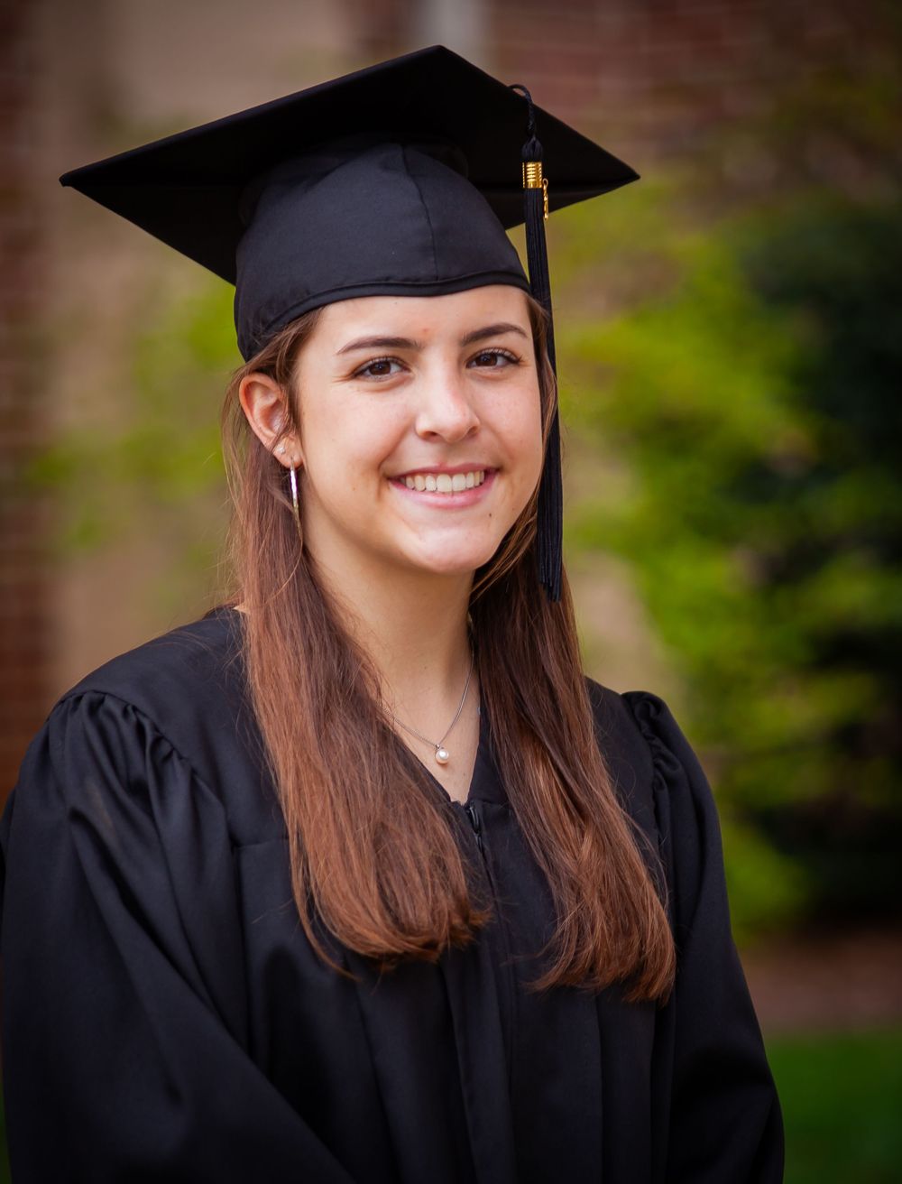 Smiling graduate in black cap and gown posing outdoors with green foliage background.