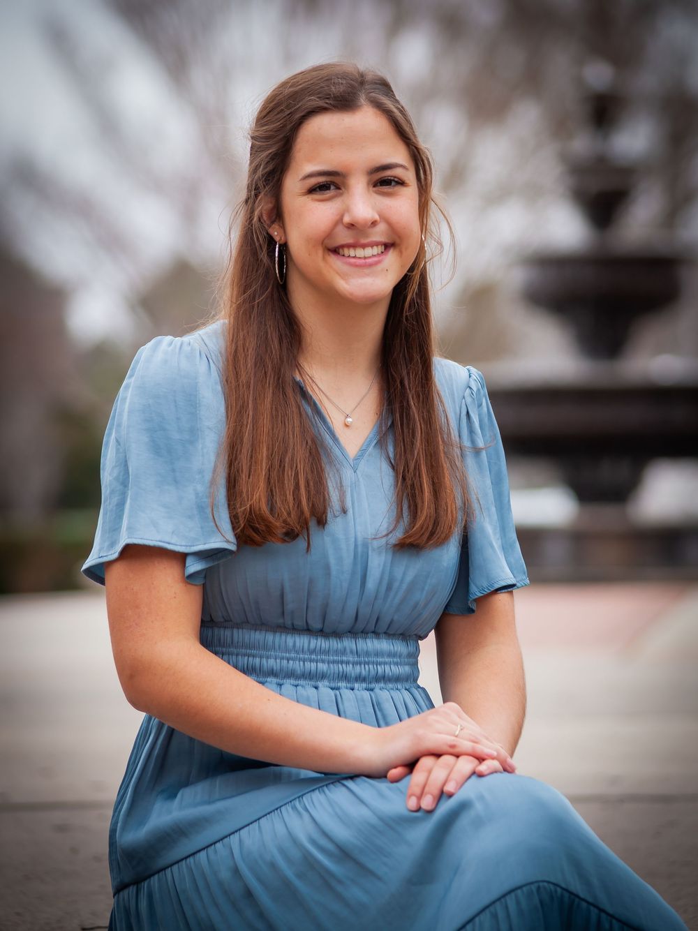 Young woman in blue dress sitting near fountain smiling for senior portrait photo.