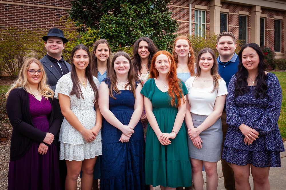 Group of smiling college students posing together outside brick building in formal attire.