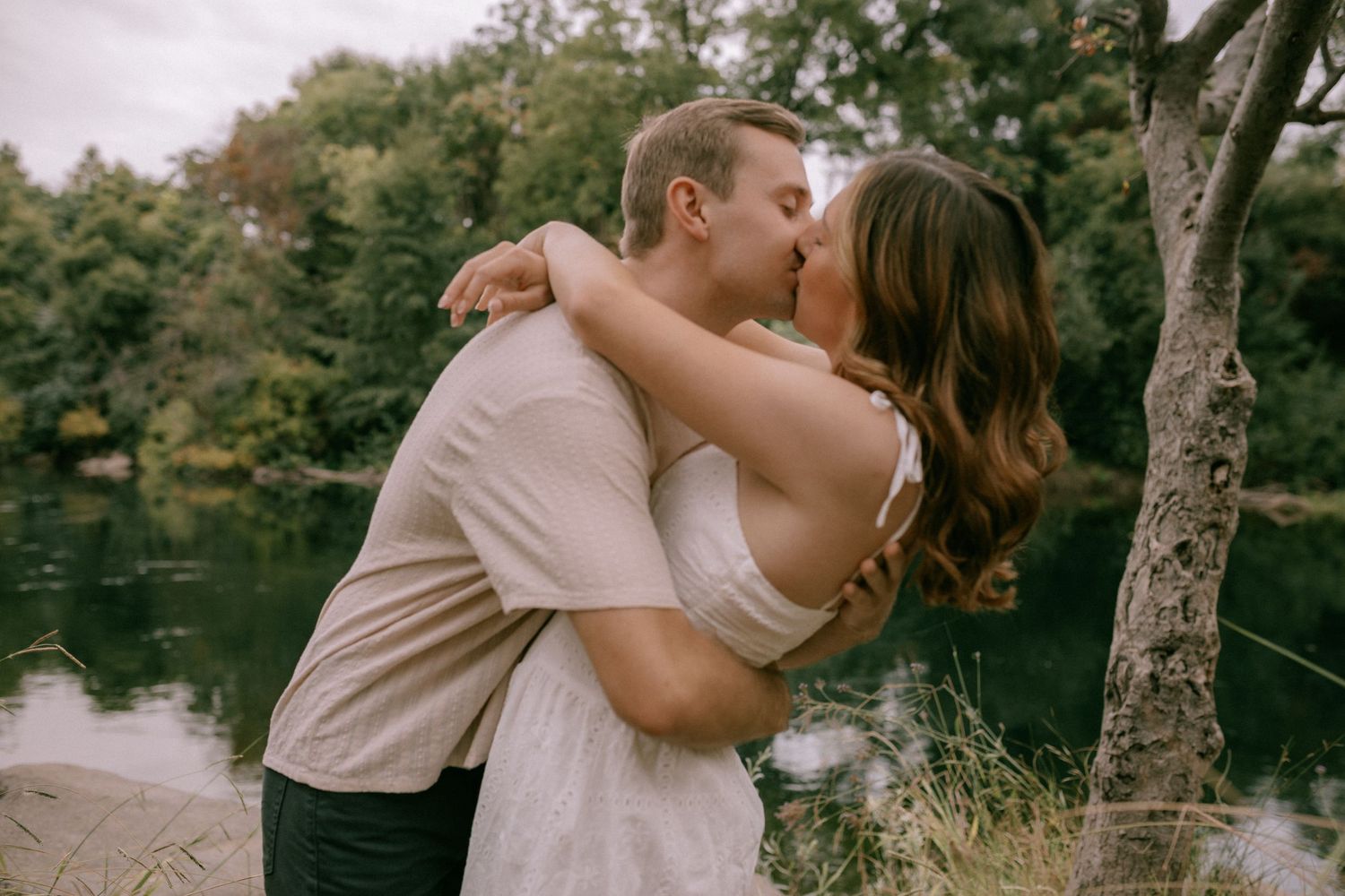 A romantic couple embraces by a serene lake surrounded by trees on a warm summer afternoon.
