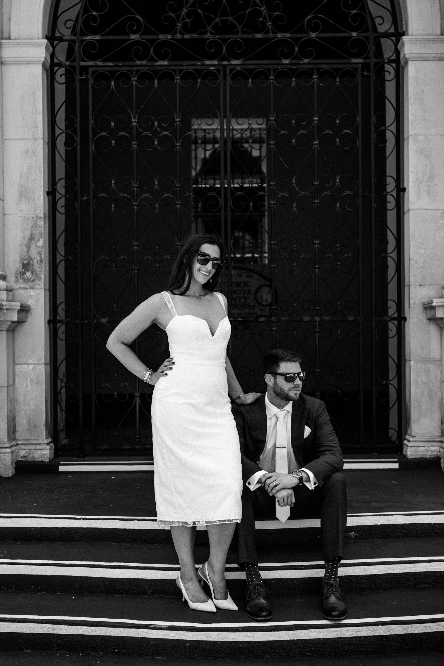 Stylish couple in black and white photo posing on stone steps with ornate iron door behind them.