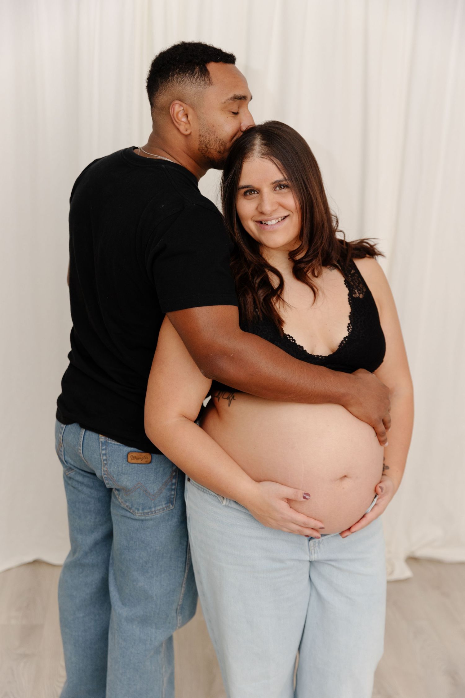 A couple shares an intimate moment in a maternity photo series showing loving embraces and belly touches against a white backdrop.