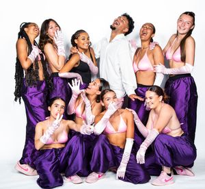 A dance group wearing purple pants and white tops poses expressively together in a dynamic studio photo.
