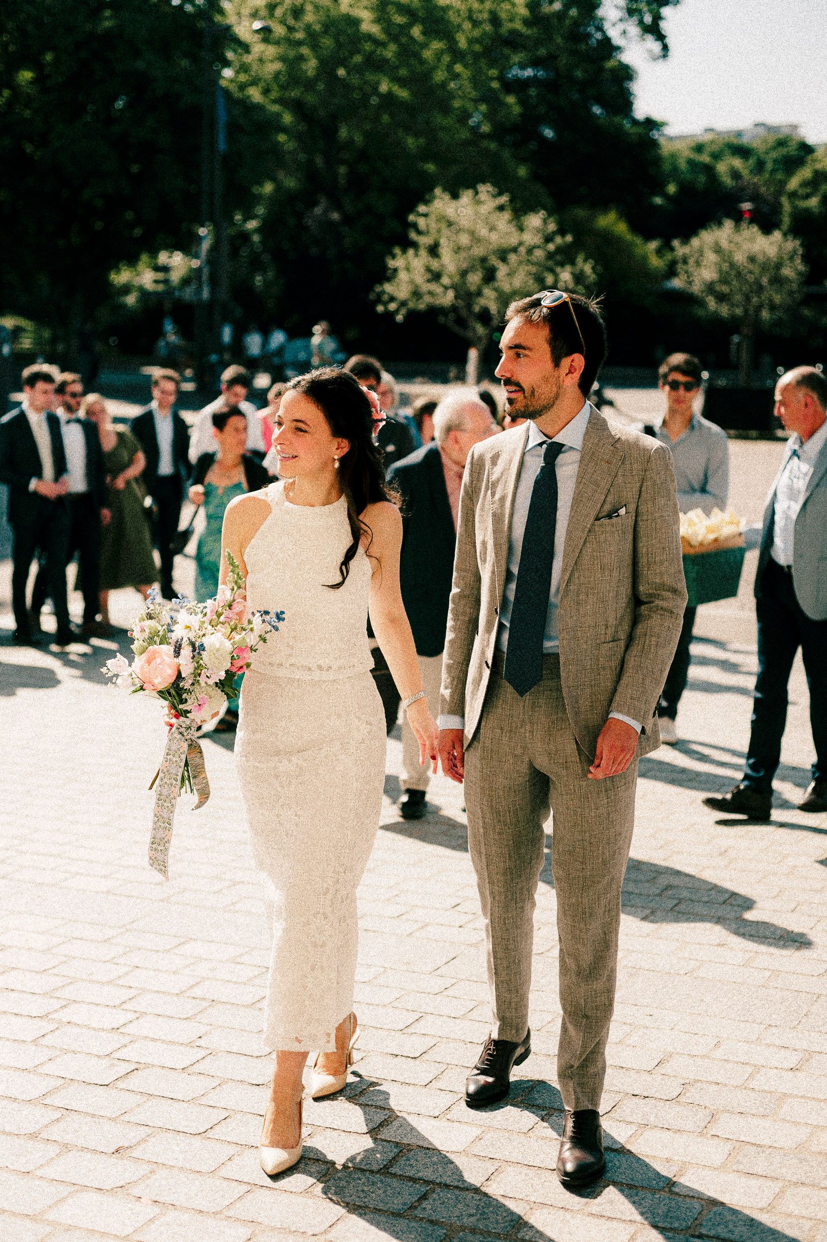Wedding party walks together on stone pavement in a line wearing white dresses and gray suits on sunny day.