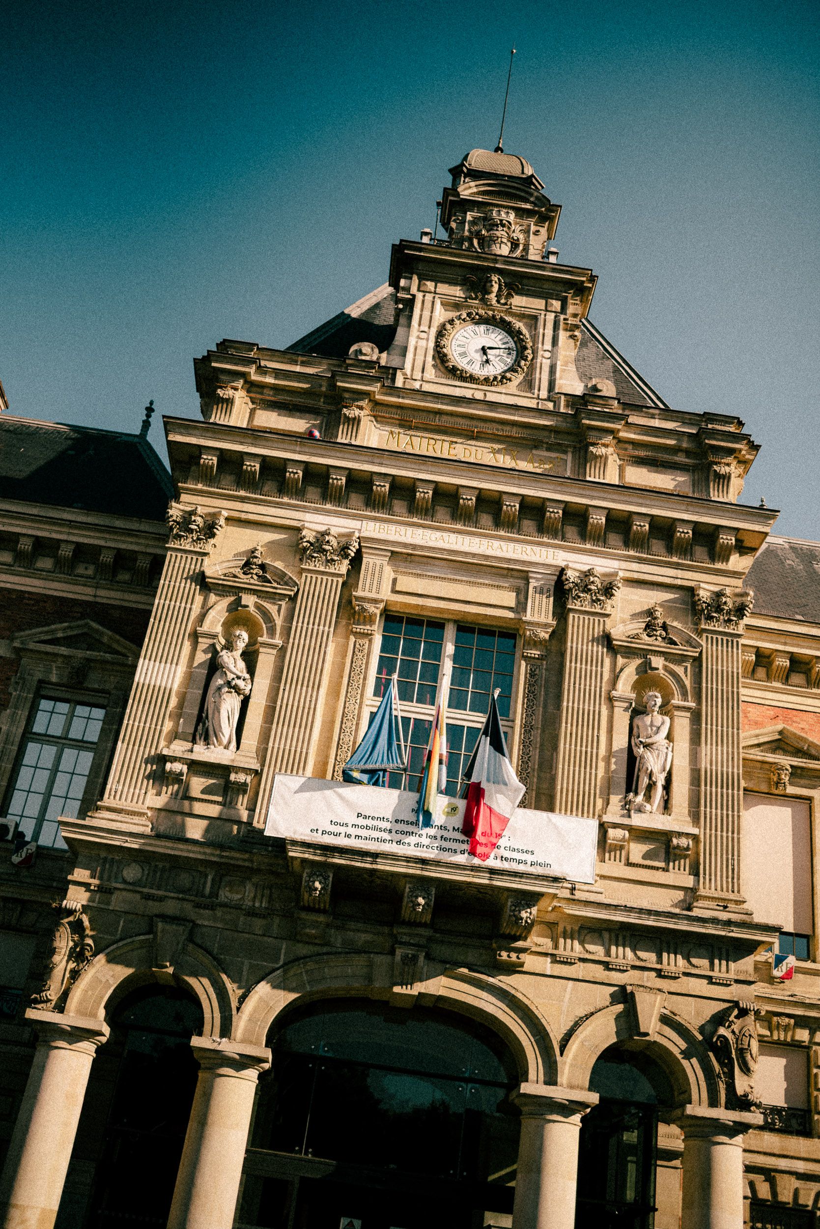 Historic European train station building with ornate classical architecture and columns against a teal-colored sky.