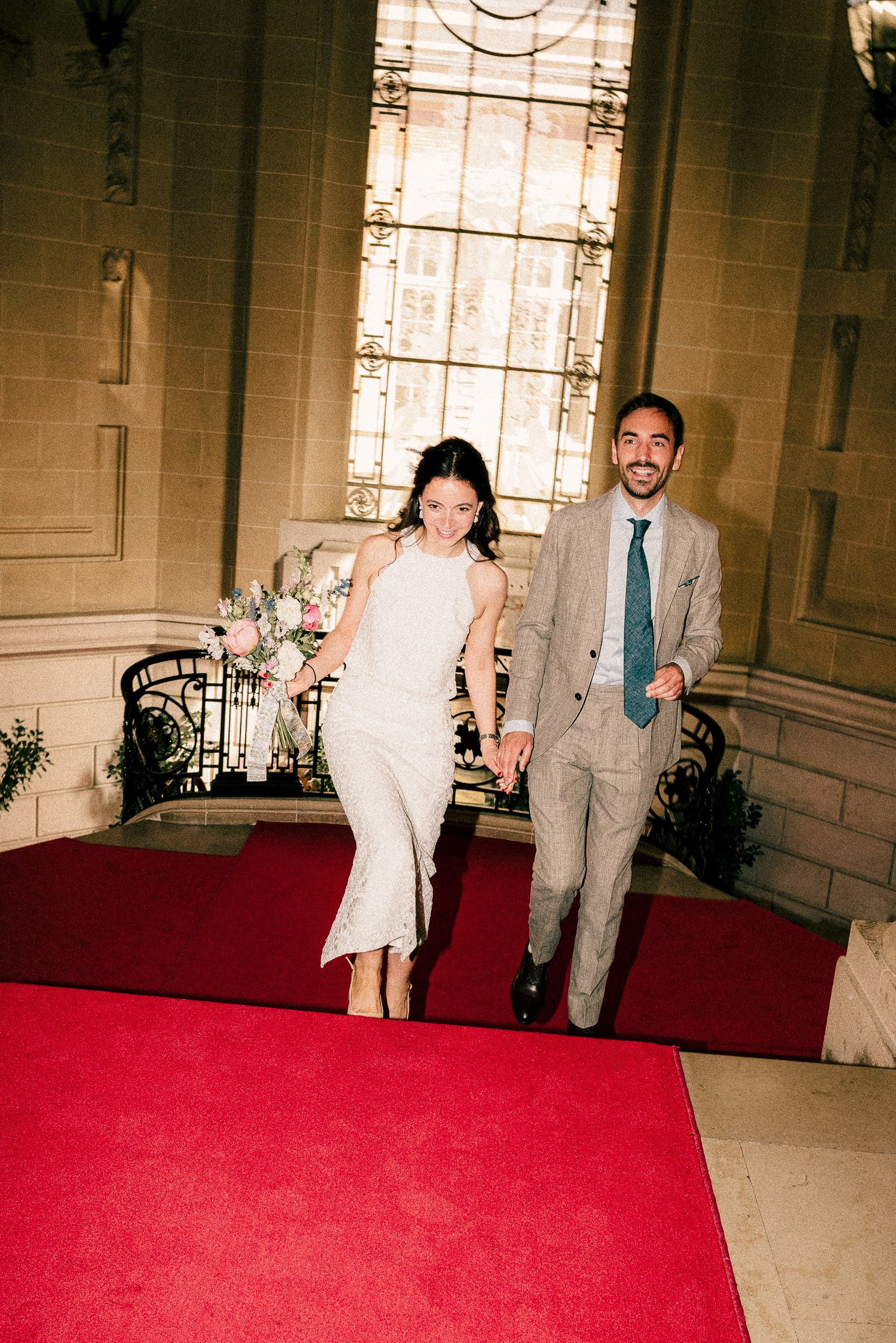A couple walks together on a red carpet in an elegant hallway with ornate architectural details.