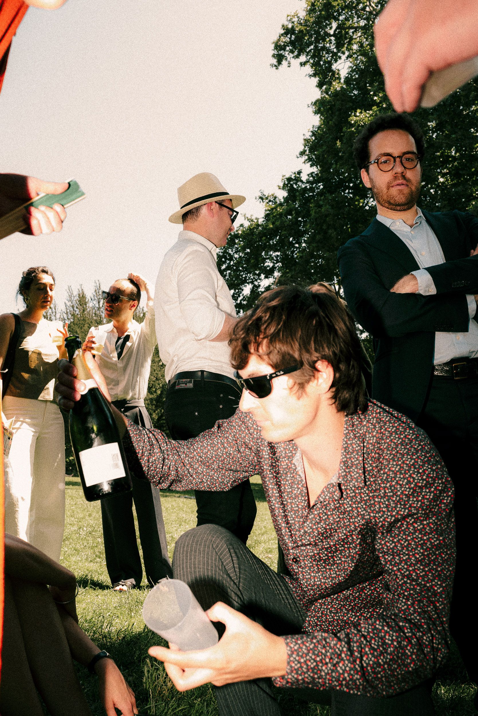 A group of people gather outdoors at a social event, with some holding drinks and wearing sunglasses on a sunny day.